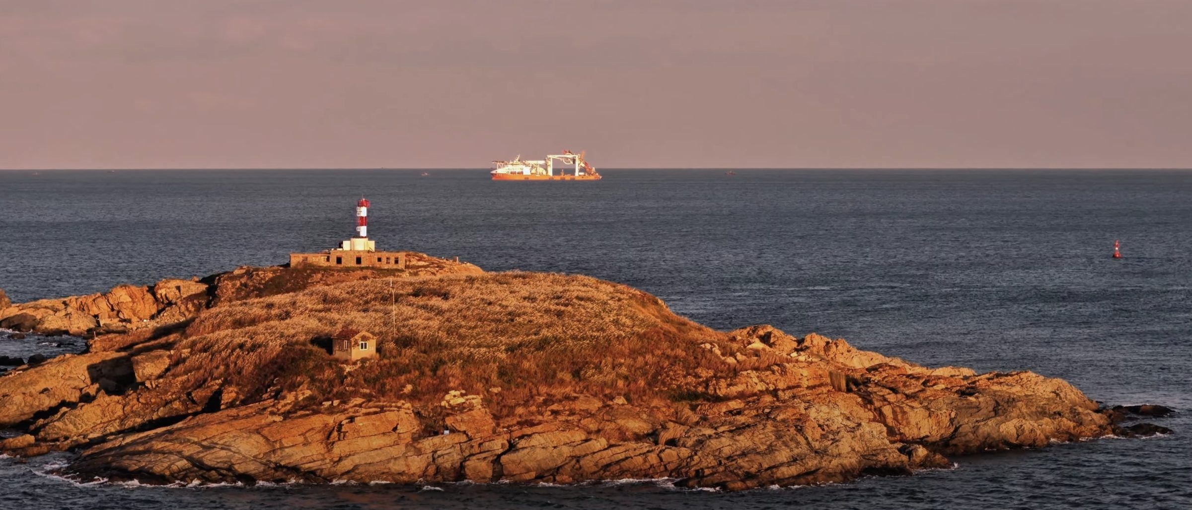 Pipelay vessel on the horizon at sunset, beyond a rocky headland with a lighthouse